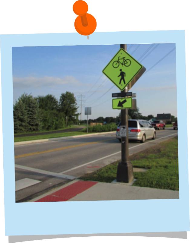 A neon yellow diamond road sign with a pedestrian and bicycle on it showing that pedestrians and bicyclists are crossing in the crosswalk. Below the sign are rectangular flashing lights.
