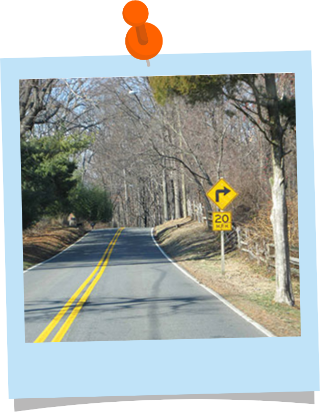 An empty two lane road through a woodsy area with a yellow diamond road sign showing a right turning arrow. Below the diamond sign is a yellow square sign that reads "20 MPH."