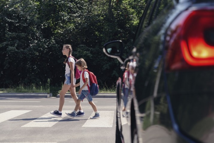 An older child and younger child crossing the street in front of a car. 