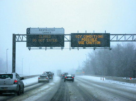 Vehicles driving on a snow-covered highway with low visibility. Overhead electronic signs display warnings: "RESTRICTED LANES CLOSED DO NOT ENTER" and "ICY CONDITIONS POSSIBLE USE CAUTION." Snow-covered trees line the sides of the road.