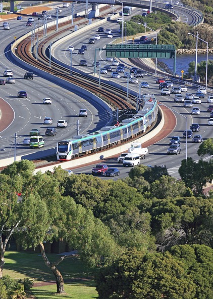 Scene of a freeway corridor with rail tracks separating the traffic traveling in five lines in each direction. Traffic is flowing well and a train is moving through the center median of the freeway. A group of green trees frames the bottom of the image.