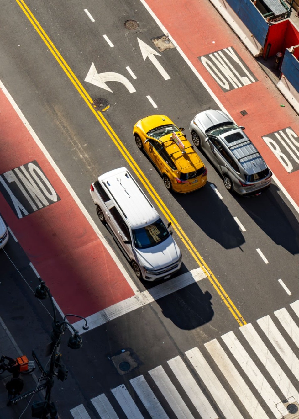 A daytime city street scene with a taxi and a silver car driving on the right side of the road. The far right lane is a bus-only lane. On the left side of the road, there are two lanes: one with a white van and another designated as a bus-only lane.
