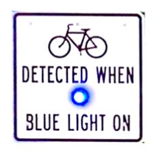 Static traffic sign reading "Bicycle detected when blue light on" with a blue light embedded in the middle. 