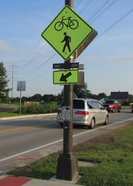 Crosswalk on a two-lane road with signs and beacons on a brown pole to warn drivers of the presence of pedestrians and bicyclists, a pushbutton-activated rectangular beacon, and a push-button to activate the beacon.