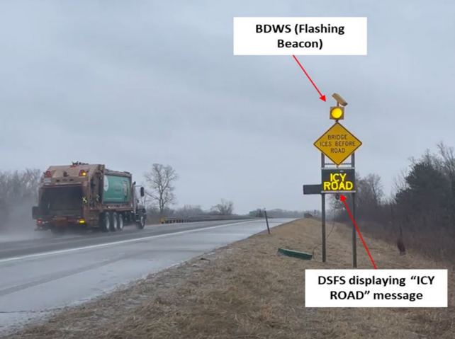 A trash truck driving on a foggy road. Off to the right side of the road sits a sign with a flashing yellow beacon on the top. Below the side there is a dynamic sign displaying a "ICY ROAD" message.