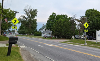 A two lane road in a suburban neighborhood. At a crosswalk there are two rectangular flashing beacons on either side of the road for pedestrian crossing.