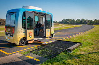 A one lane road in the middle of a field. An automated shuttle sits on the road with it's doors open and a ramp deployed.
