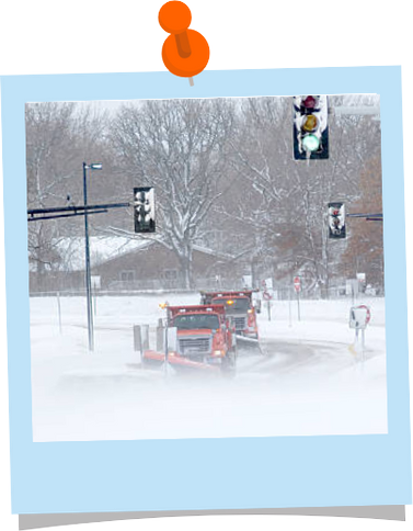 Two red snowplows driving on a road with traffic lights, pushing snow off the roads.