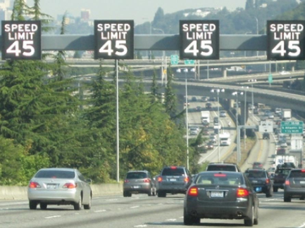 A busy multi-lane highway with vehicles traveling under a set of overhead electronic signs displaying a speed limit of 45 mph. The highway is surrounded by green trees, and multiple overpasses are visible in the background. Directional signs indicate exits for W Seattle and Columbian Way. Traffic is moderate, with cars in all lanes.