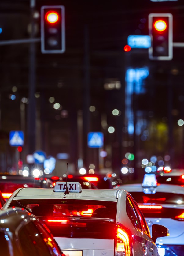 A nighttime city street scene featuring a line of taxis waiting at a traffic light. The cars' taillights create a bright red glow, and blurred lights from traffic signals and street lamps illuminate the background, adding to the bustling urban atmosphere.
