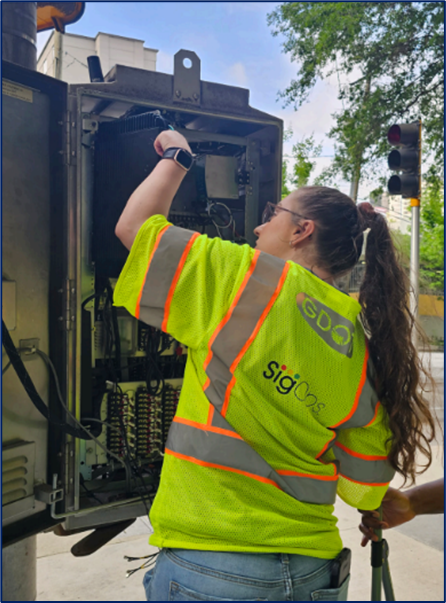 A worker wearing a safety vest installing a device at controller cabinet.